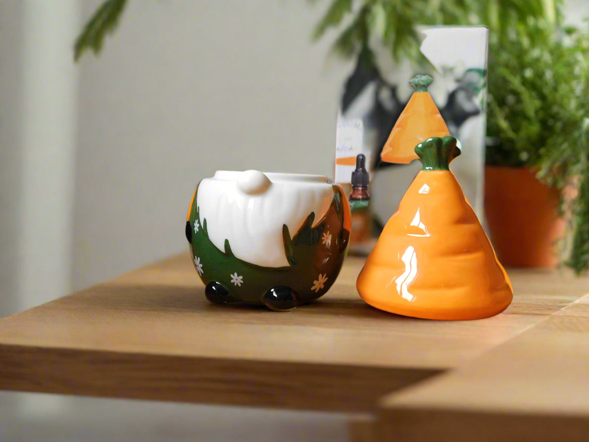 A ceramic oil/wax burner in the shape of a gnome / gonk with a carrot patch on the hat, placed on a wooden shelf. view from the front with the hut on the top of the counter top