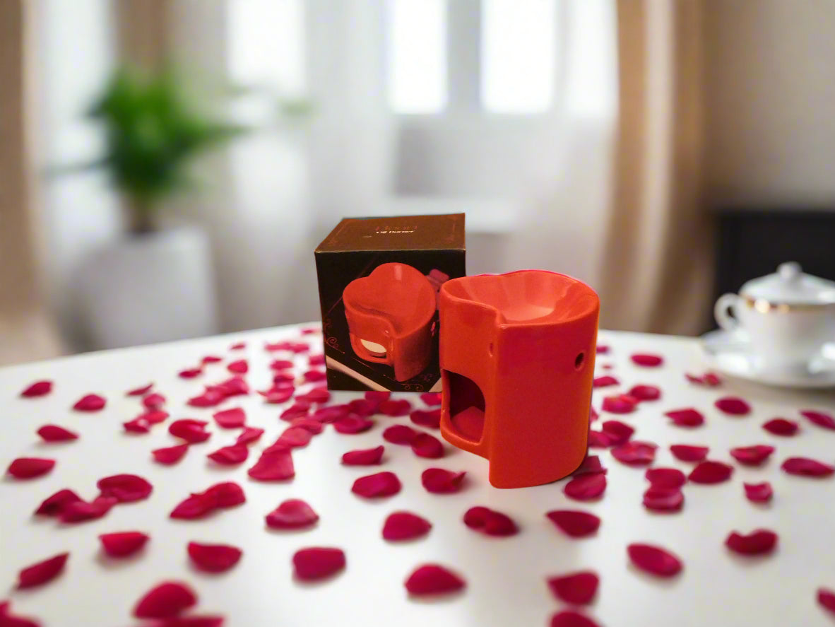 A red heart-shaped oil burner with scattered rose petals and a lit candle in the background. View from the side