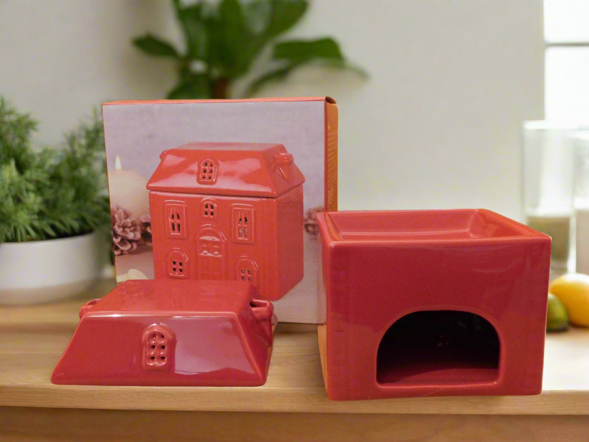 A red ceramic wax burner designed to look like a house, displayed on a wooden surface with a photo of the product and some decorative items in the background. View from the back