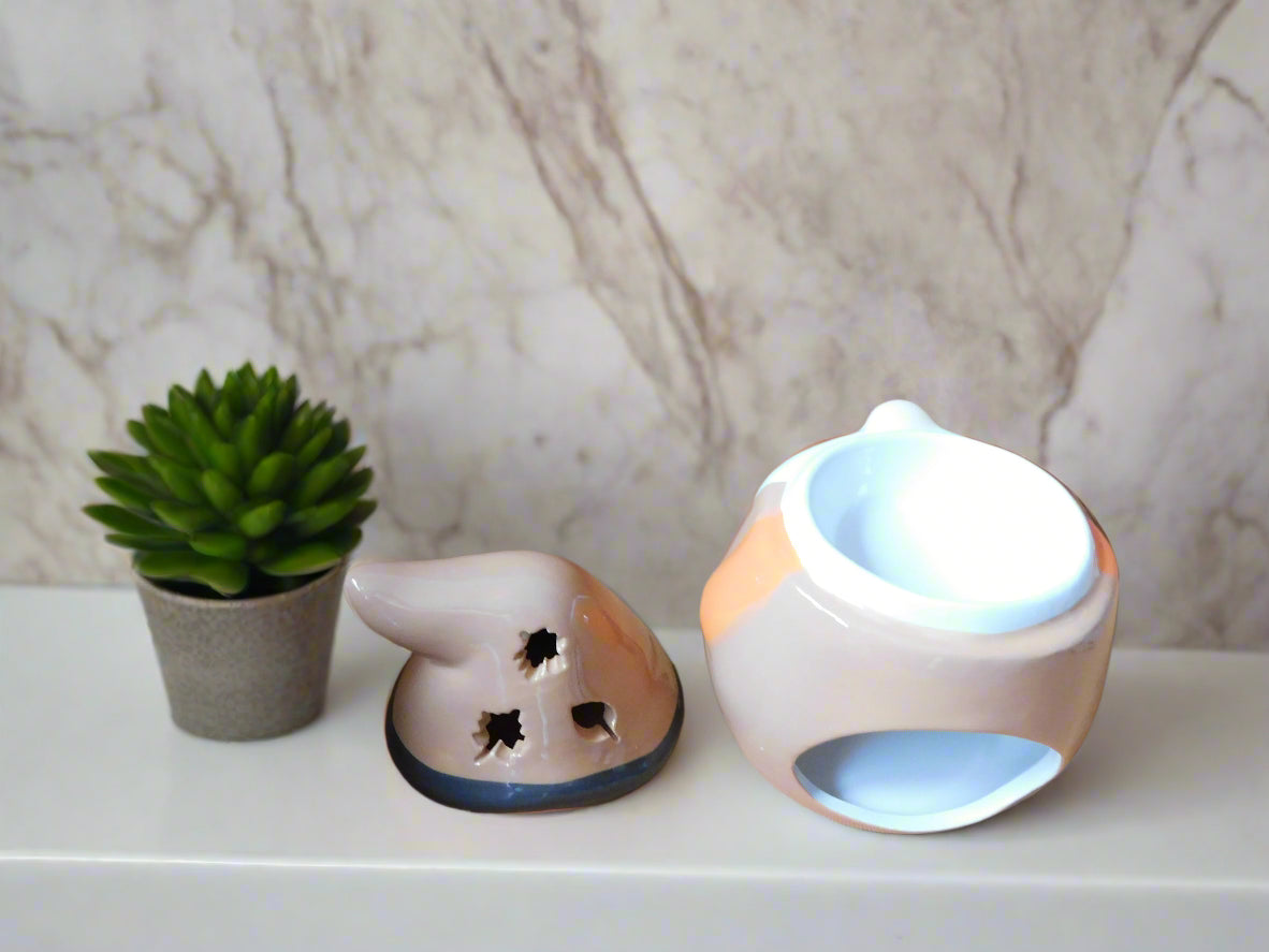Ceramic oil/wax burner with a design of a Gonk, in orange and brown colors, placed on a white surface with a plant to its left. The hut of the gonk is on the top of the counter. View from the back