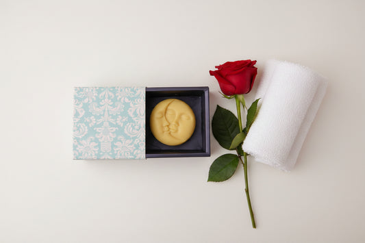 A pale yellow crescent moon shaped soap bar resting inside an open dark box next to its light blue and white damask-patterned lid