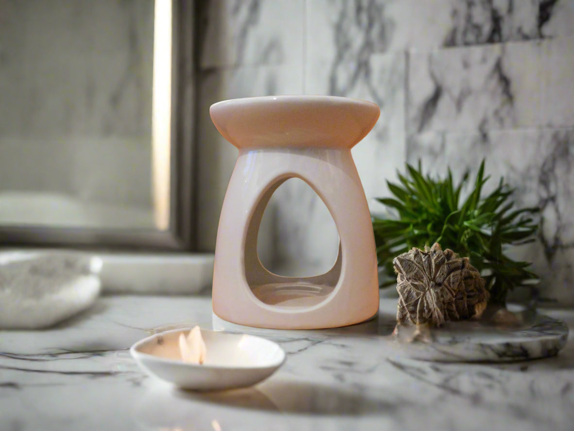 A white ceramic wax burner on a marble surface with a plant in the background and a lit candle on a dish to the front.