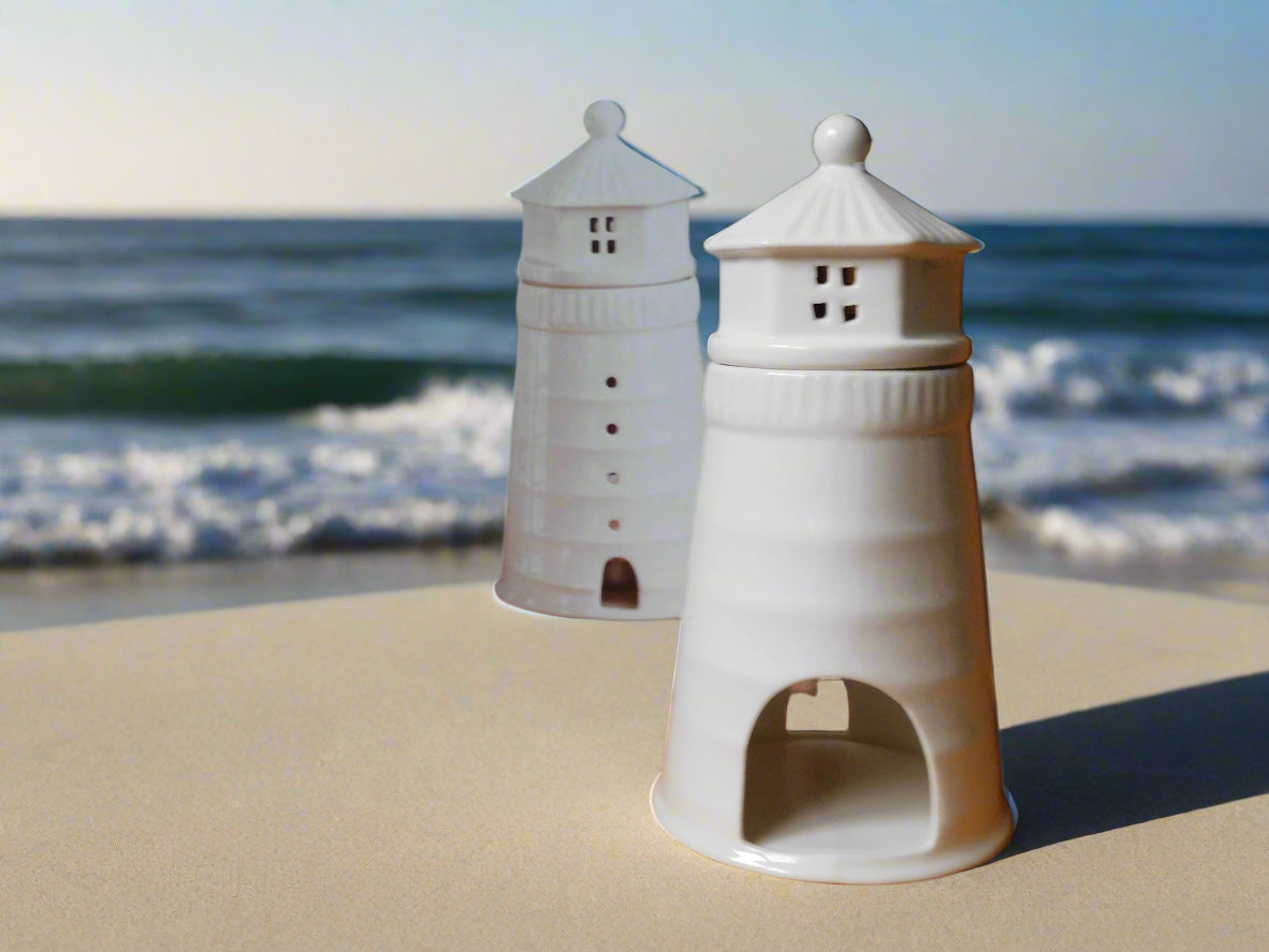 A white ceramic oil and wax burner designed to resemble a lighthouse, displayed on a beach with the ocean in the background. View from the back