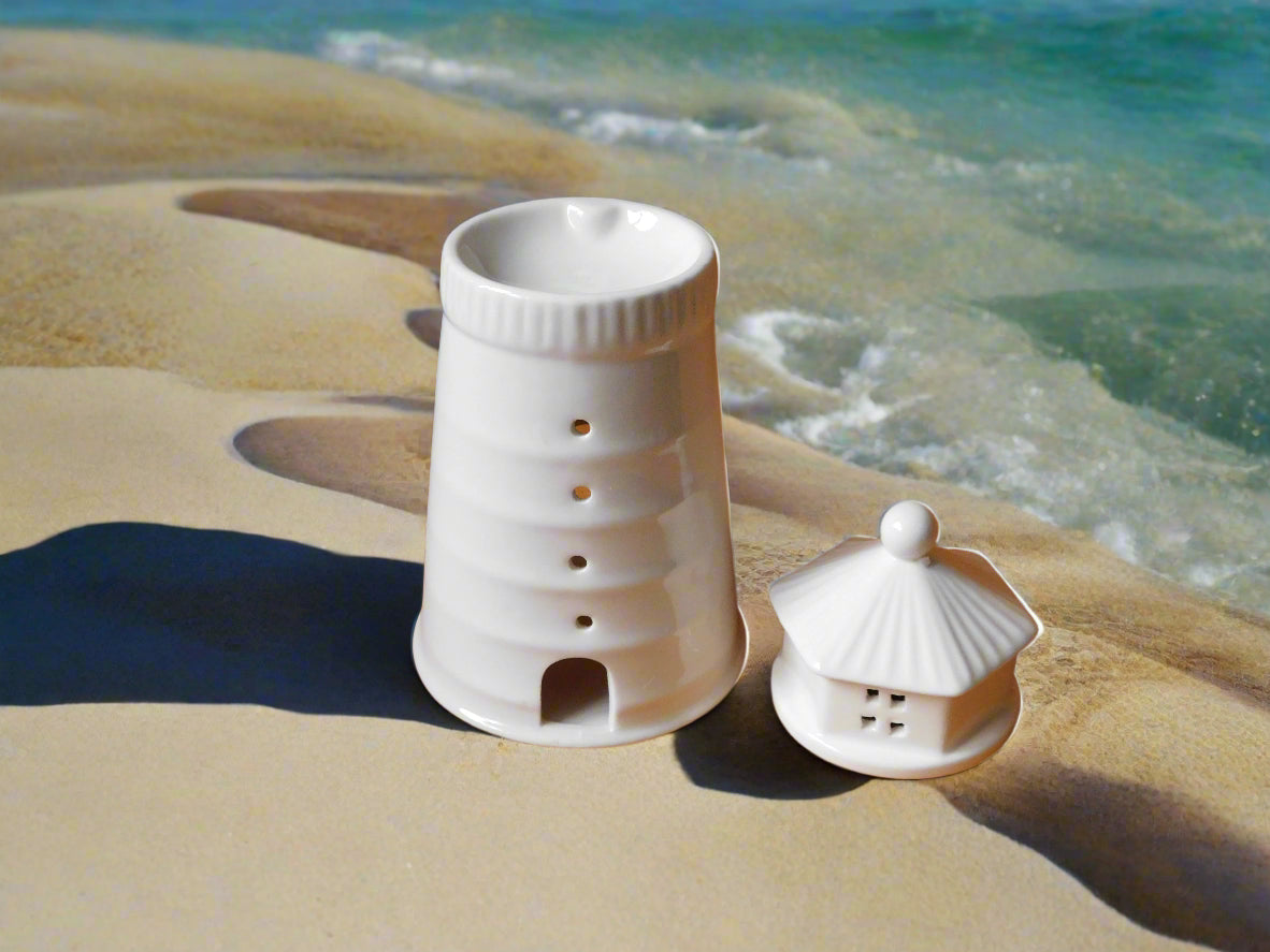 A white ceramic oil and wax burner designed to resemble a lighthouse, displayed on a beach with the ocean in the background. The top is on the top of the sand.