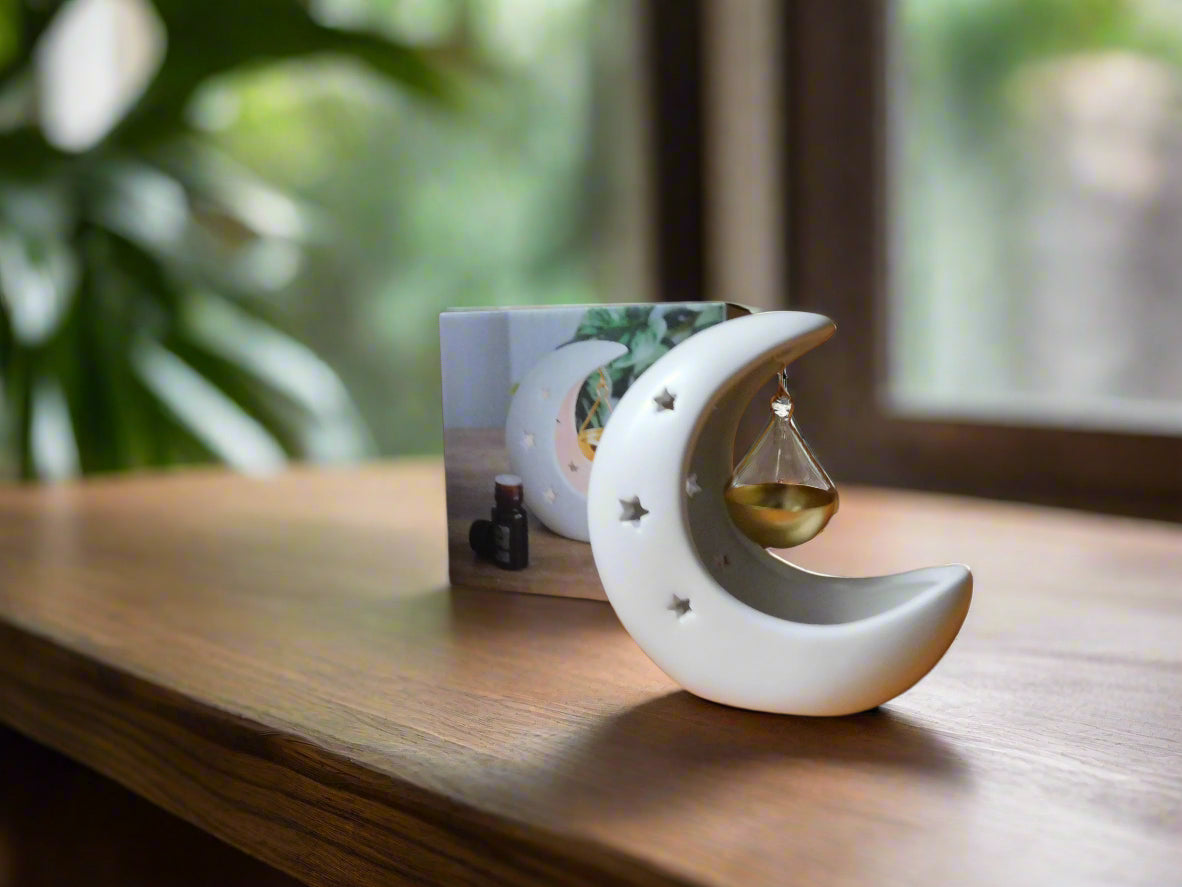 A white crescent moon-shaped ceramic oil/wax burner with a gold dish hanging by a string, displayed on a wooden surface with a blurred greenery background.