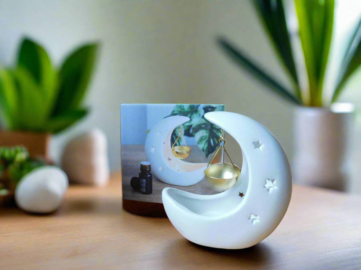 A white crescent moon-shaped ceramic oil/wax burner with a gold dish hanging by a string, displayed on a wooden surface with a blurred greenery background.View from the back
