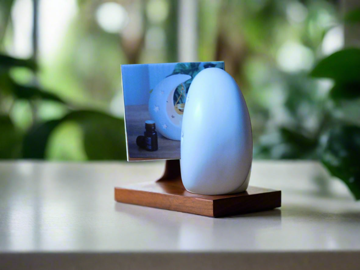 A white crescent moon-shaped ceramic oil/wax burner with a gold dish hanging by a string, displayed on a wooden surface with a blurred greenery background. View from the side