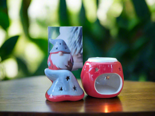 A red ceramic wax burner designed to resemble a gonk, with a white beard and grey hat, placed on a wooden surface with foliage in the background. View from the back.