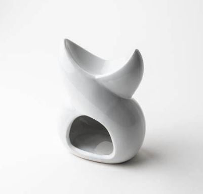 A white ceramic wax melt burner with a twisted design, placed on a wooden surface with a plant and other home decor items in the background. View from the side