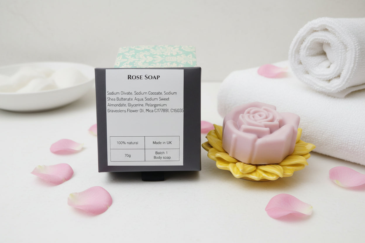 A bar of rose soap displayed next to its packaging box on an orange textured surface. The soap is shaped like a pink rose blooming from a yellow sunflower, and the box features a white label with product details.
