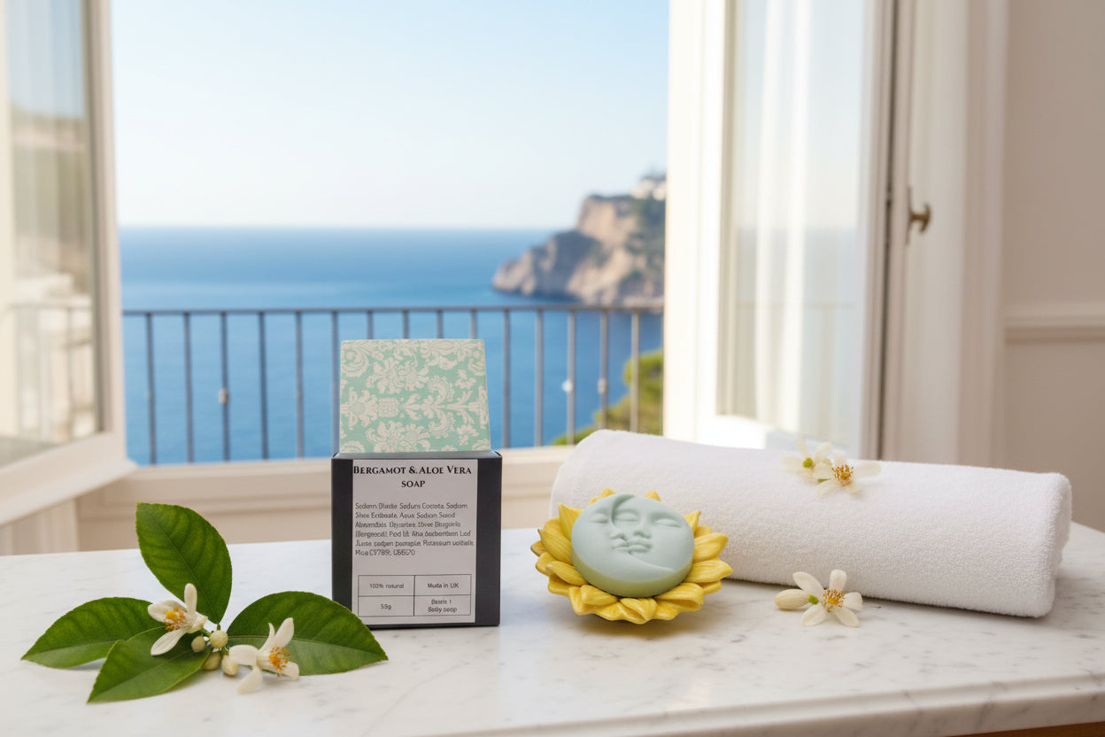 Bergamot & Aloe Vera soap bar displayed next to its packaging box on a textured orange-brown background. The soap is a pale sage green with a carved face, set inside a yellow sunflower-shaped holder, while the box features a light green damask pattern and a dark gray base with white text.