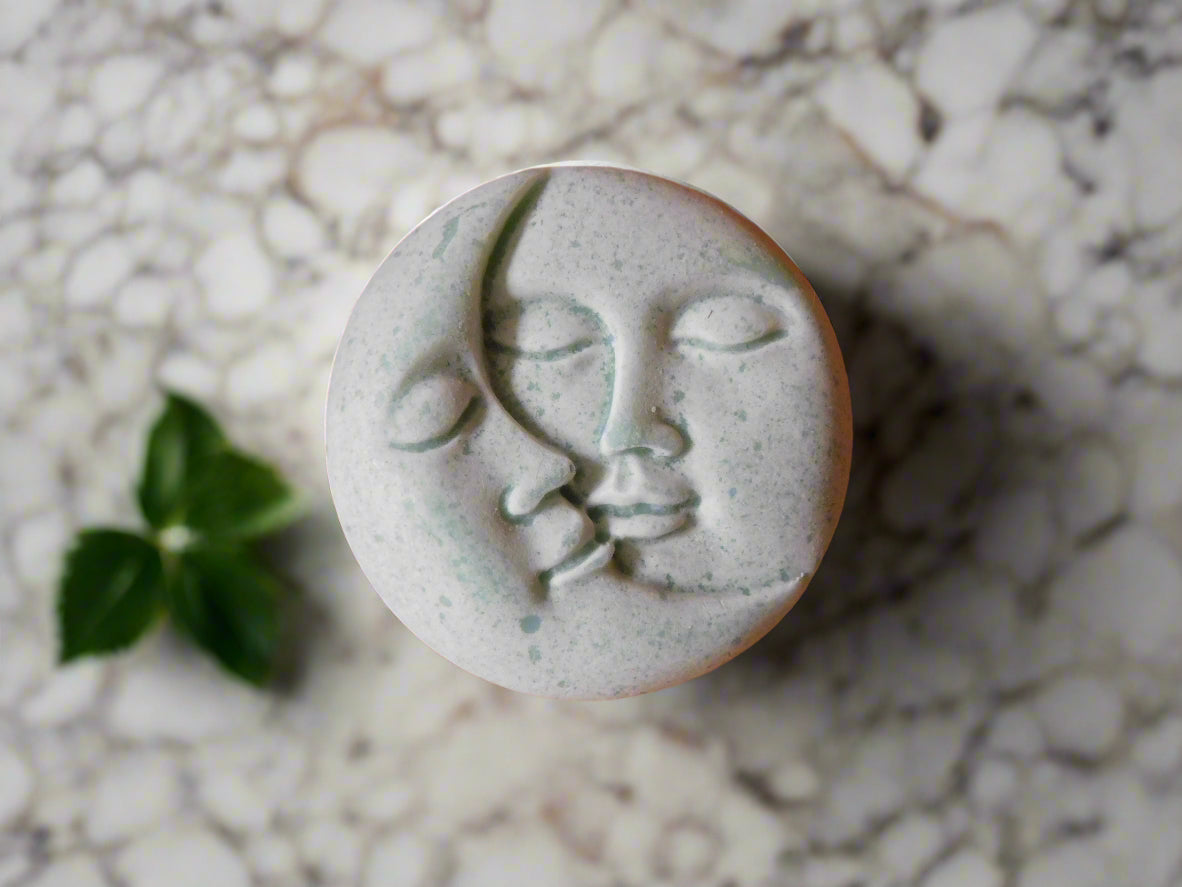 Handmade round soap with a design resembling two faces embracing, placed on a marble surface with a small green leaf to the side.