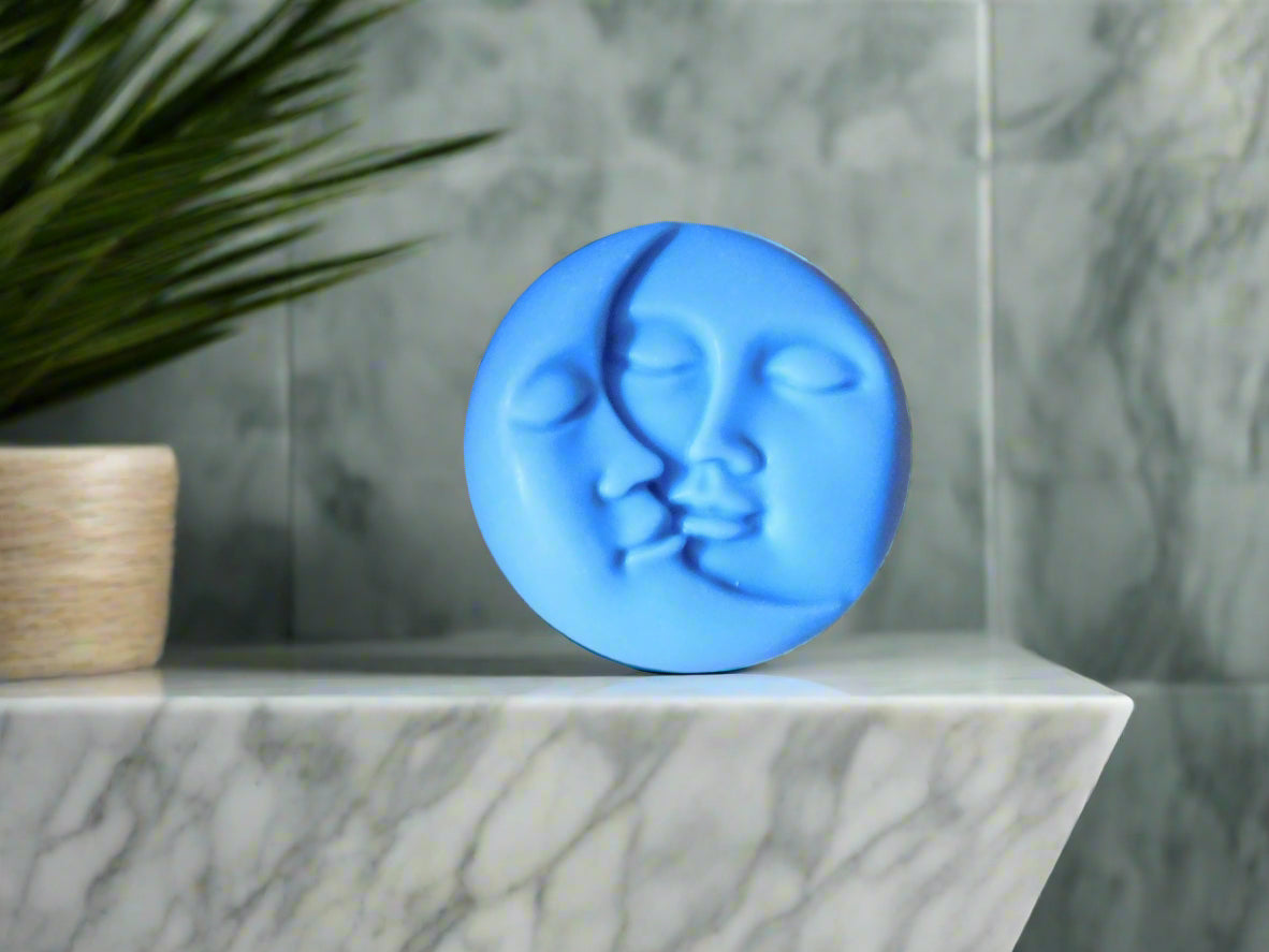 A handmade bar of soap with a blue color and a design resembling a half moon face on it, placed on a marble surface.