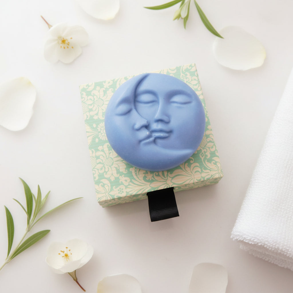 A round blue soap bar with a sculpted face merging with a crescent moon, displayed on a square patterned backing card