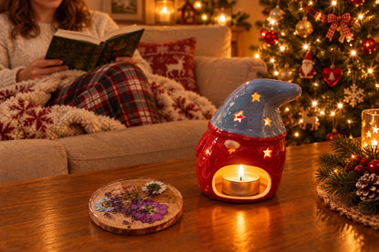 A ceramic Christmas gnome warmer and matching decorative piece displayed next to its product box on a tan leather sofa. The gnome features a light blue body with star-shaped cut outs and a red sleeve, while the accompanying round piece contains dried purple and white flowers under clear resin