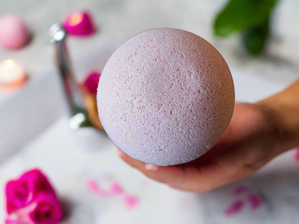 Pink bath bomb situated on the top of a hand with roses all around the bath that it trying to put into the bath 