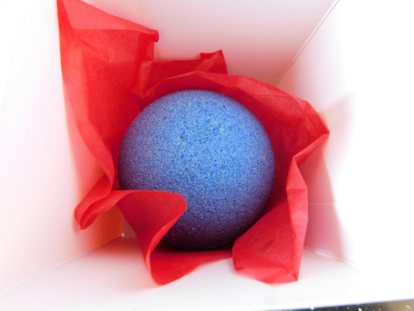 Blue bath bomb wrapped in red tissue paper inside a box on a white background