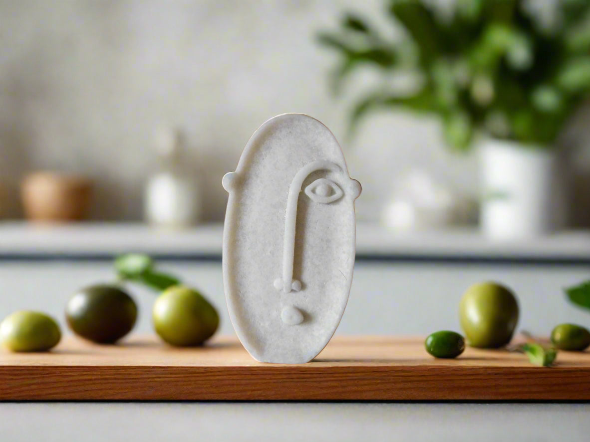 A bar of soap with a geometric oval face design on a shelf with plants and olives in the background.