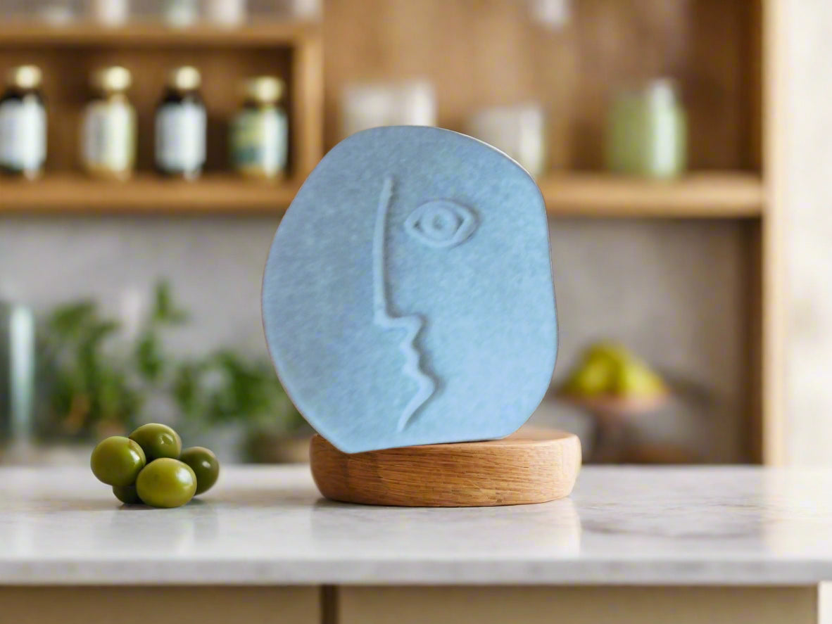 A bar of soap with a geometric face design on a shelf with olives in the background.