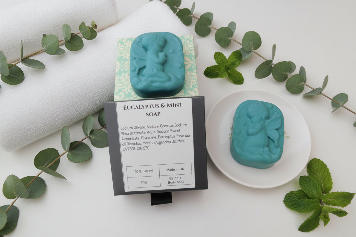 A dark green body soap bar with an embossed figure, shown both wrapped in patterned paper and standing upright next to a white dish with a soap bar. A black label card reads 'EUCALYPTUS & MINT SOAP' with ingredients and product details