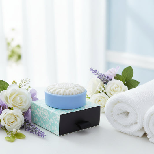 A round white soap bar with a sunflower-like embossed top, resting on a light blue and white patterned square box with a black ribbon pull
