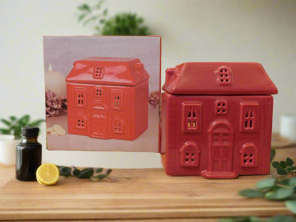 A red ceramic wax burner designed to look like a house, displayed on a wooden surface with a photo of the product and some decorative items in the background.