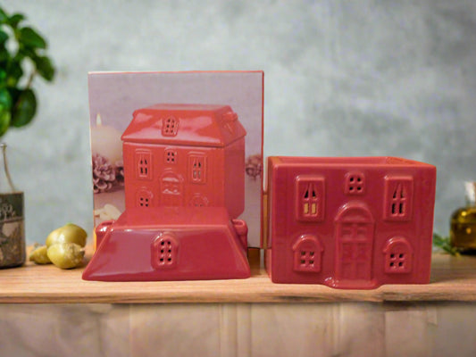 A red ceramic wax burner designed to look like a house, displayed on a wooden surface with a photo of the product and some decorative items in the background. The roof of the house it is on the top of the wooden surface