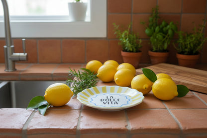 La dolce vita tricket dish. Colour yellow and blue with black ink written in the middle of the dish LA DOLCE VITA in capital letters. There are lemons all around the dish on the top a mediterranean counter top