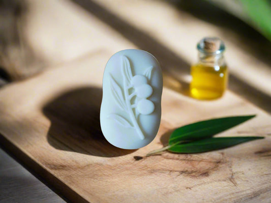 A handmade Lily soap with an olive design, placed on a wooden surface with a small bottle of oil and a leaf in the background.
