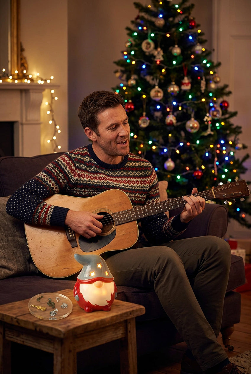 A glowing red gonk sits atop the table beside a musical coaster. A man on the sofa strums his guitar, singing carols, while a Christmas tree stands in the background.