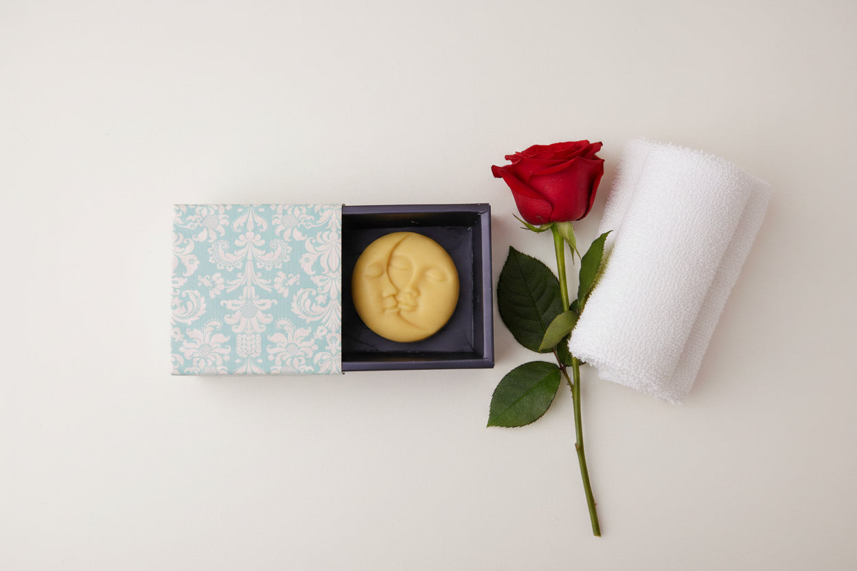 A pale yellow crescent moon shaped soap bar resting inside an open dark box next to its light blue and white damask-patterned lid