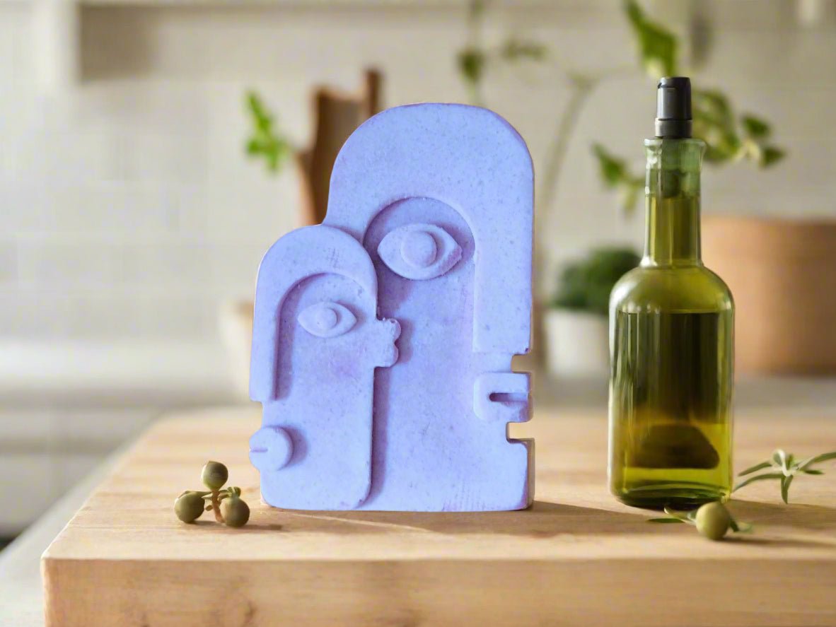 A bar of soap with a geometric face design on a shelf with plants in the background olive oil bottle on the right handside and olives in both side of the soap