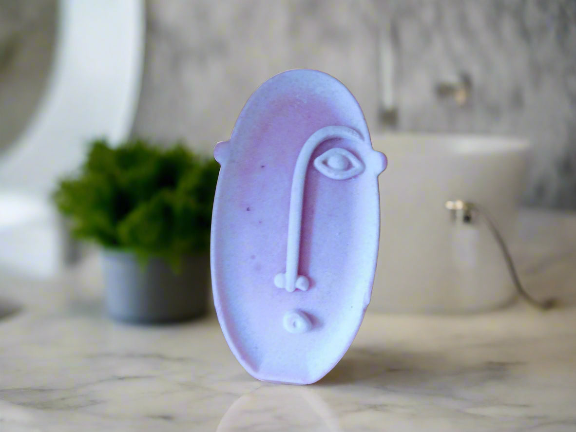 A bar of soap with a geometric face design on a marble table top with plants in the background and a sink