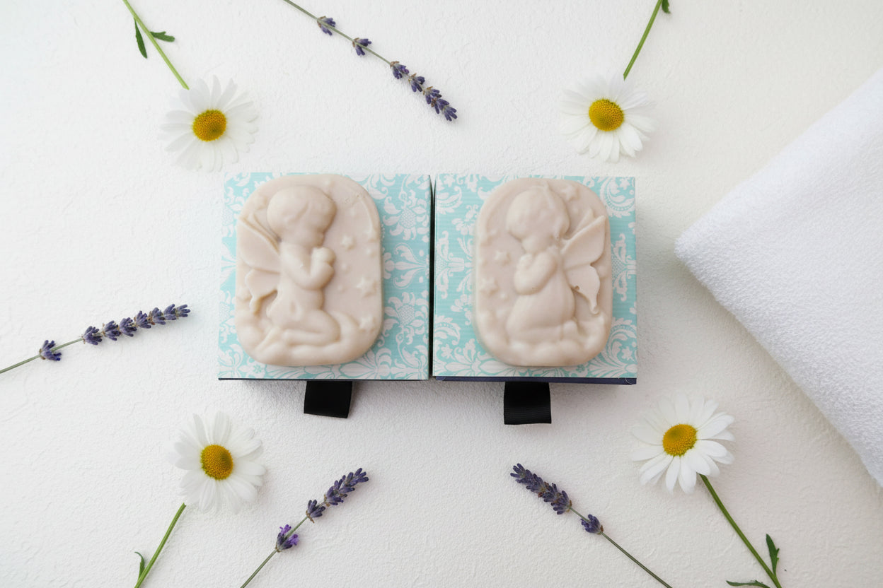 Two rectangular soap bars with embossed cherub designs, displayed side-by-side on a light blue and white patterned