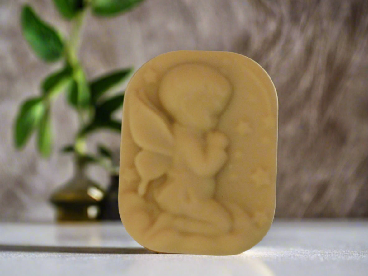 A handmade bar of soap with an embossed boy angel shape design on top, placed on a counter with soft lighting in the background.