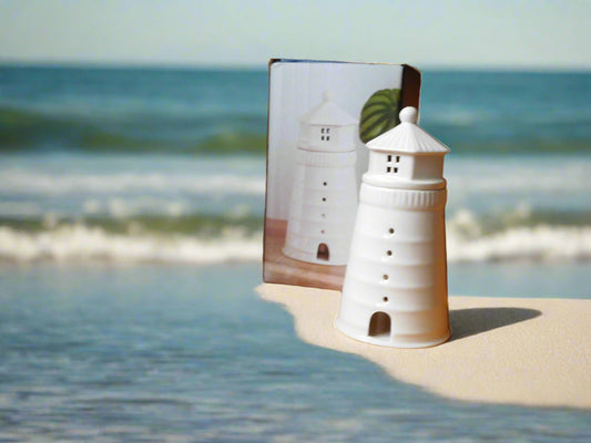 A white ceramic oil and wax burner designed to resemble a lighthouse, displayed on a beach with the ocean in the background. 