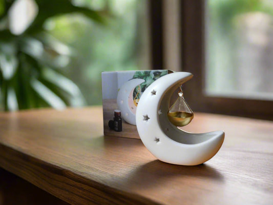 A white crescent moon-shaped ceramic oil/wax burner with a gold dish hanging by a string, displayed on a wooden surface with a blurred greenery background.