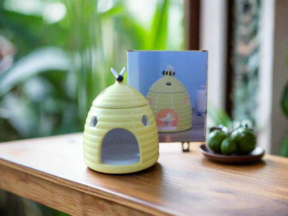 A yellow ceramic beehive oil burner displayed on a wooden surface with a plant background. The burner is designed to look like a beehive with a top lid.