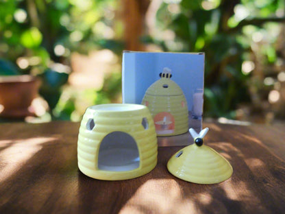 A yellow ceramic beehive oil burner displayed on a wooden surface with a plant background. The burner is designed to look like a beehive with a top lid that it is situated on the top of the surface. View from the back.