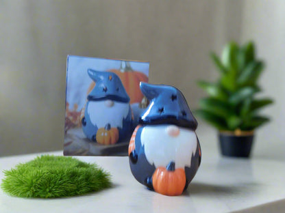 A grey ceramic wax burner designed to look like a gnome / gonk, placed on a white surface with a green moss-like mat to its left and a picture depicting a similar scene in the background.