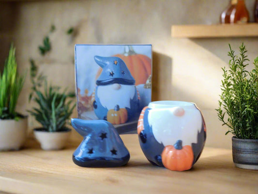 A grey ceramic wax burner designed to look like a gnome / gonk, placed on a white surface with a green moss-like mat to its left and a picture depicting a similar scene in the background. The hut is on the top of the counter