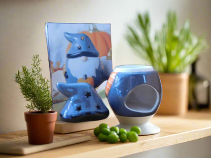 A grey ceramic wax burner designed to look like a gnome / gonk, placed on a white surface with a green moss-like mat to its left and a picture depicting a similar scene in the background. View from the back. The hut it is on the top of the counter