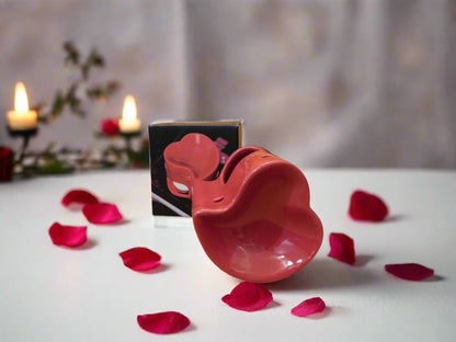 A red heart-shaped oil burner with scattered rose petals and a lit candle in the background. View from the front