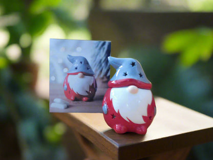 A red ceramic wax burner designed to resemble a gonk, with a white beard and grey hat, placed on a wooden surface with foliage in the background.