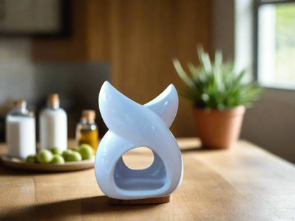 A white ceramic wax melt burner with a twisted design, placed on a wooden surface with a plant and other home decor items in the background.