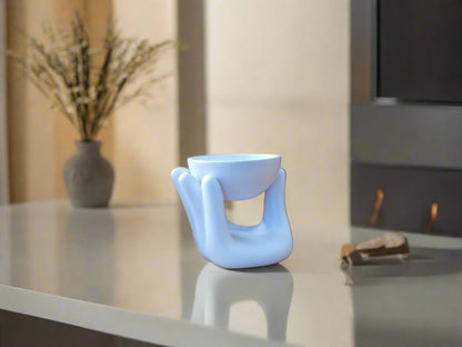 A white ceramic oil burner designed in the shape of a hand, sitting on a table with a bowl in the center.